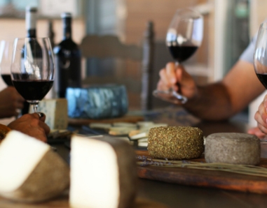 escena en una mesa con varias personas que brindan con una copa de vino tinto y comen quesos artesanos de diferentes tipos