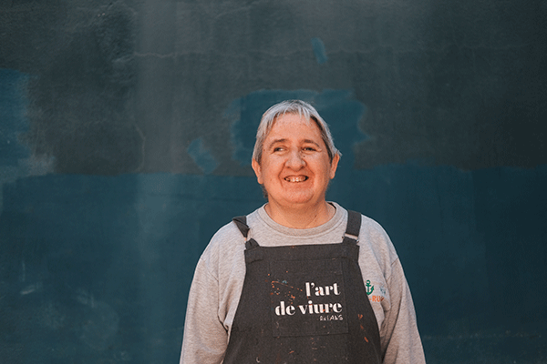 Persona con delantal de "L'Art de Viure" de AMPANS sonriendo frente a un fondo azul oscuro.